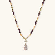 Gold necklace with iridescent baroque pearl pendant, purple amethyst beads, and crystal star charm on cream background